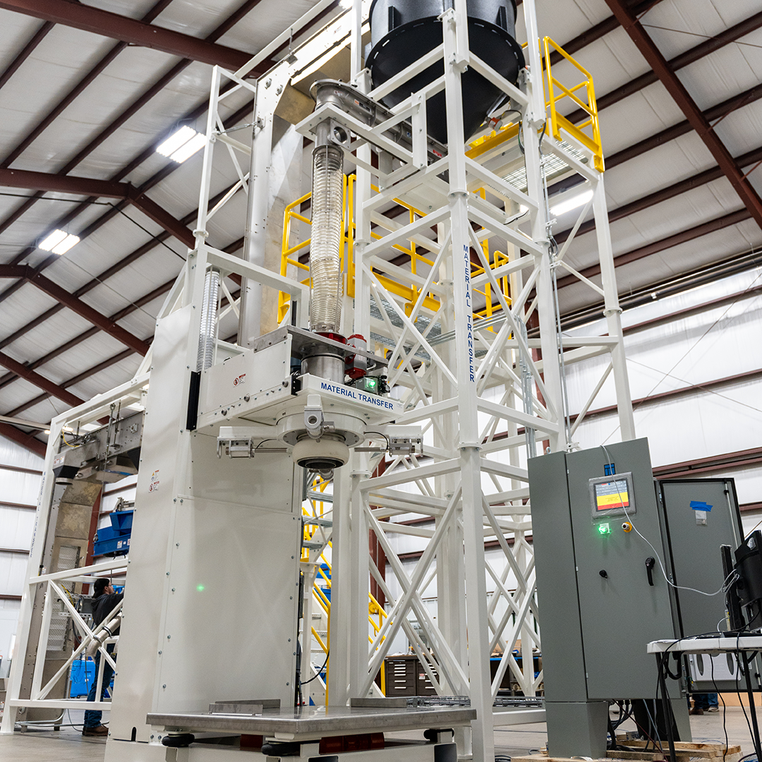 The image showcases a large industrial setup for bulk bag filling, designed and manufactured by Material Transfer & Storage. The system includes structural frames, conveyors, and a control panel, with a focus on precision and efficiency. The prominent "Material Transfer" branding emphasizes the company's expertise in bulk material handling solutions. The setup features integrated components such as a filling spout, support structure, and control mechanisms, suitable for a variety of material handling applications.